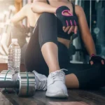 Mujer descansando en el gimnasio junto a pesas y botella de agua recuperandose post ejercicio